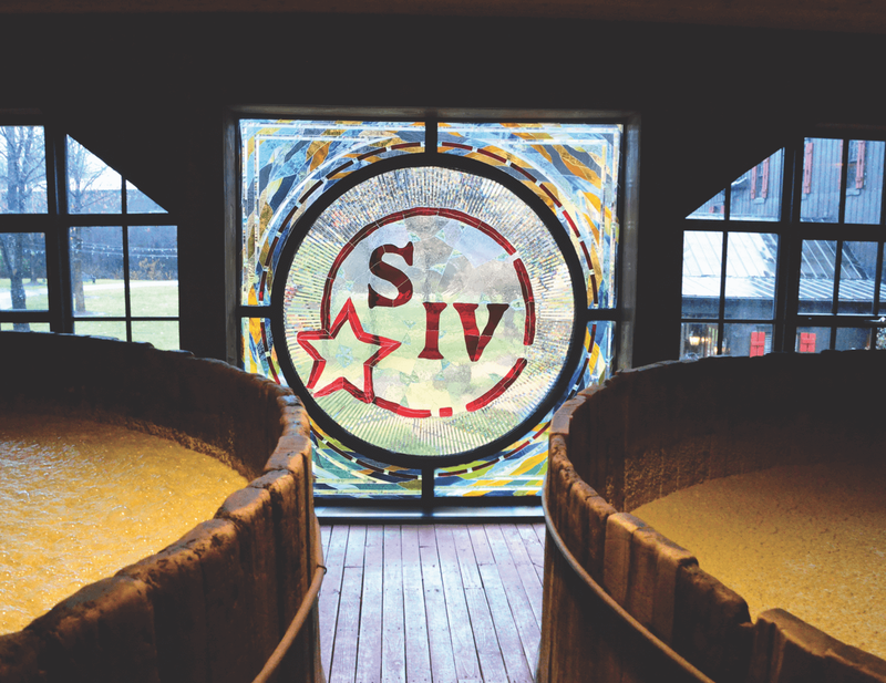 A custom stained glass window with the Maker's Mark logo is pictured between two barrels of aging whiskey